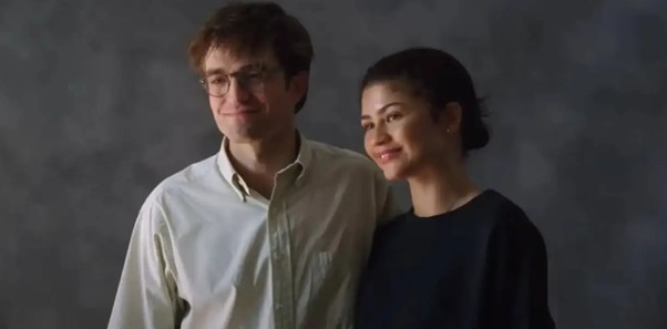 Un homme et une femme souriant ensemble devant un fond texturé, l'homme porte une chemise claire et des lunettes, tandis que la femme porte un pull noir.